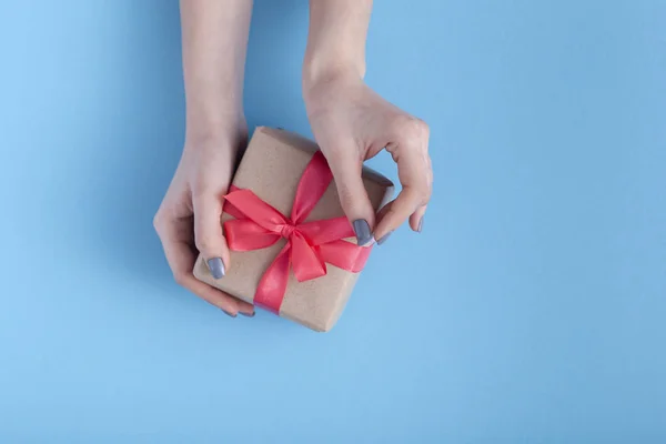 Niña sosteniendo un regalo en las manos, mujeres con caja de regalo con un lazo de cinta roja atada en las manos sobre un fondo azul pastel, vista superior, concepto de vacaciones, amor y cuidado — Foto de Stock