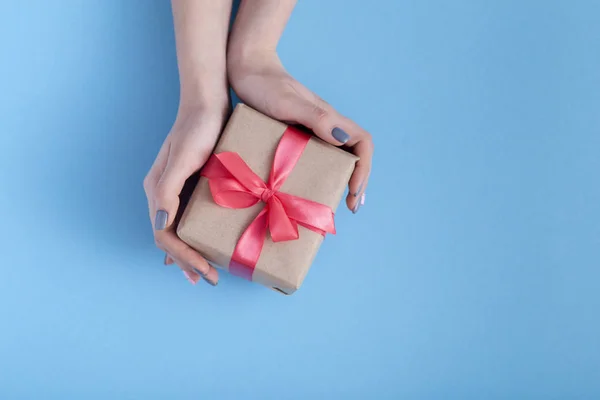 Dziewczyna trzyma prezent w ręce, kobiety z pudełka prezent z wiązanej czerwoną wstążką łuk w ręce na pastelowe kolorowe niebieskie tło, widok z góry, koncepcja wakacje, miłość i opieka — Zdjęcie stockowe
