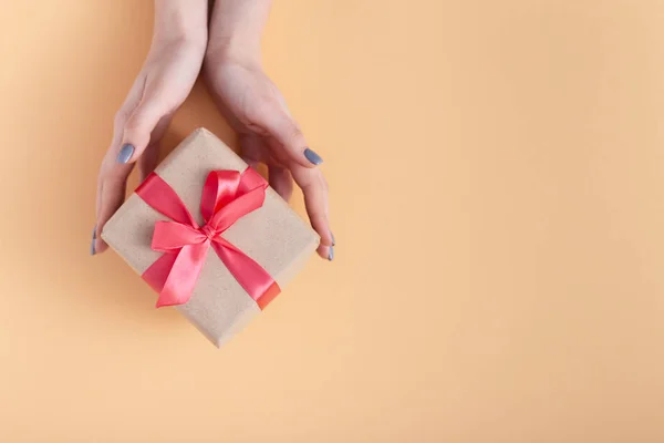 Menina segurando um presente em mãos, mulheres com caixa de presente em mãos envolvidas em papel artesanal decorativo com um laço de fita vermelha amarrada em um fundo de cor pastel, vista superior, férias conceito, amor e cuidado — Fotografia de Stock