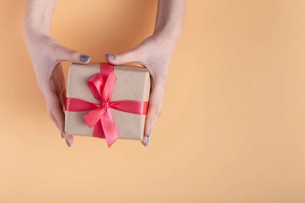 Menina segurando um presente em mãos, mulheres com caixa de presente em mãos envolvidas em papel artesanal decorativo com um laço de fita vermelha amarrada em um fundo de cor pastel, vista superior, férias conceito, amor e cuidado — Fotografia de Stock