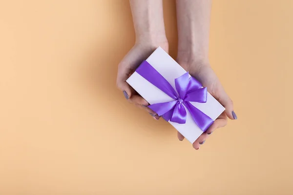 Niña sosteniendo un regalo en las manos, las mujeres con caja de regalo con un lazo de cinta lila atada en las manos sobre un fondo naranja de color pastel, vista superior, concepto de vacaciones, amor y cuidado — Foto de Stock