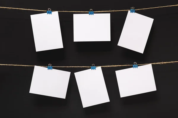 empty paper cards attached with colorful clothespins on linen rope on a black background, blank templates concept office and photo industry, decor