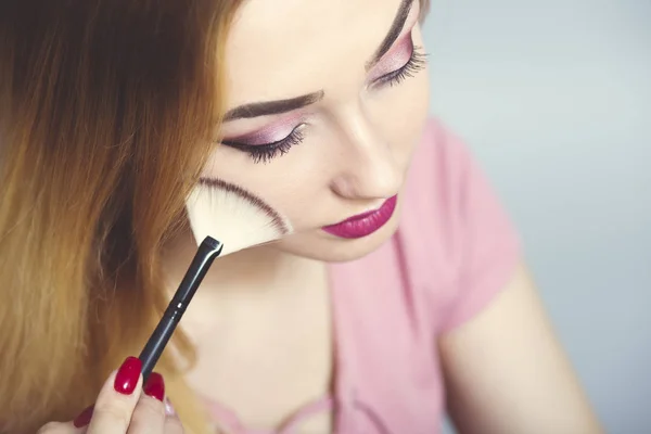 Porträt eines schönen jungen Mädchens beim Make-up in einem Taschenspiegel in einem Atelier, eine Frau, die ihr Gesicht mit einem Pinsel abstaubt, Kosmetik- und Schönheitskonzept — Stockfoto