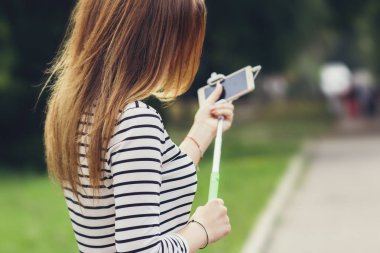 bir selfie çubuğu kullanarak bir akıllı telefon üzerinde bir şehir parkı bir sosyal ağ için bir fotoğraf yapma kız