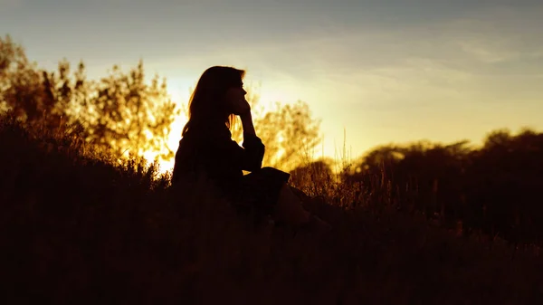 Silhouette of a young woman sitting on a hill at sunset, figure of girl in the autumn landscape — Stock Photo, Image