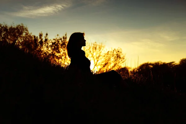 Silhouette of a young woman sitting on a hill at sunset, figure of girl in the autumn landscape — Stock Photo, Image