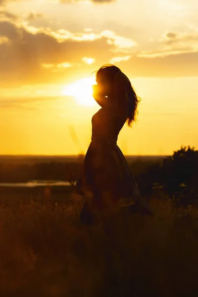 Silhueta de uma menina bonita em um vestido ao pôr do sol em um campo, figura de mulher jovem apreciando a natureza, conceito de lazer — Fotografia de Stock