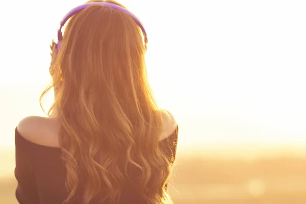 自然の音楽を聴くヘッドフォンでティーンエイジャーの女の子は、若い女性がリラックスして、夏のフィールドで夕日を見ています — ストック写真