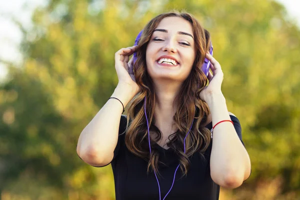 Porträtt av en vacker lycklig tonåring flicka med hörlurar på huvudet, ung kvinna lyssnar på musik på naturen i fält — Stockfoto