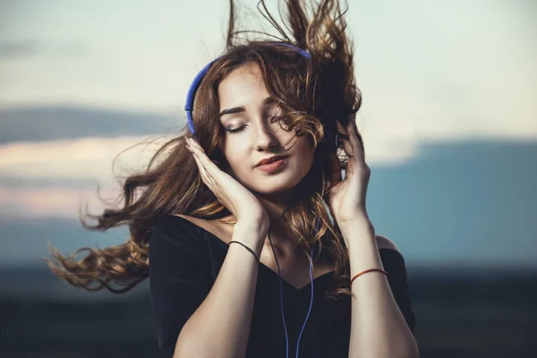 Romantiskt porträtt av en vacker flicka i hörlurar med flygande hår från vinden, ung kvinna lyssnar på musik utomhus — Stockfoto