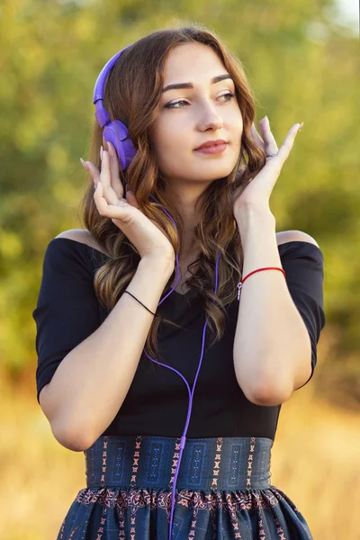 Porträtt av en vacker flicka i hörlurar på huvudet, ung kvinna lyssnar på musik på naturen i fält — Stockfoto
