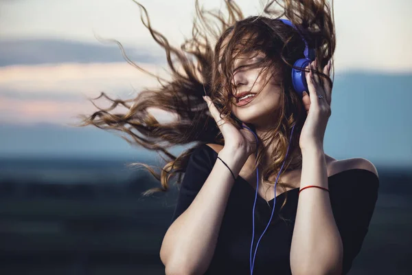 Porträtt av en vacker flicka i hörlurar med flygande hår från vinden, ung kvinna lyssnar på musik på naturen i fält — Stockfoto