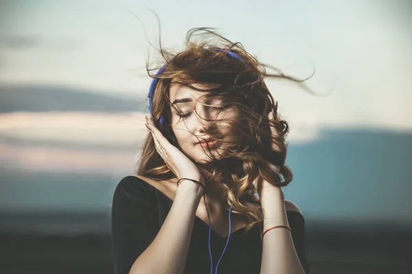 Romantiskt porträtt av en vacker flicka i hörlurar med flygande hår från vinden, ung kvinna lyssnar på musik utomhus — Stockfoto