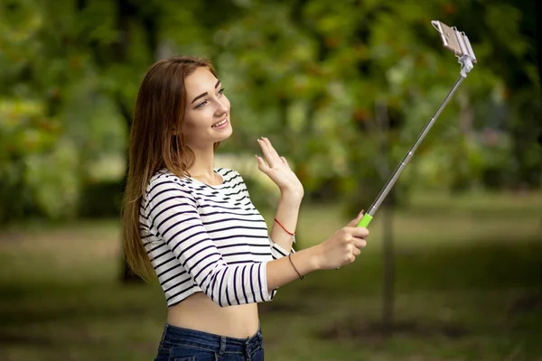 Fille heureuse communiquer avec des amis sur Internet et salue en agitant la main dans un parc de la ville avec smartphone sur bâton selfie, concept de style de vie adolescent — Photo