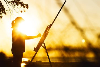 bir şelon üzerinde tuval üzerine boyalar ile bir resim boyama bir kadın silueti, gün batımında bir alanda doğa üzerinde sanat yapan boya fırçası ve paletli kız