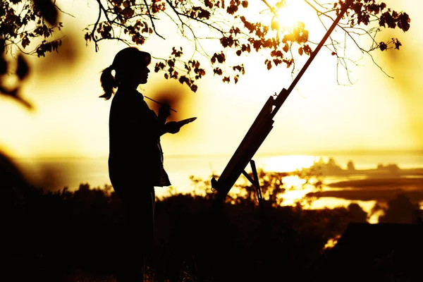 Silhueta de uma jovem mulher desenhando um quadro em um cavalete na natureza, fêmea em pé sob a árvore com pincel e paleta de artista envolvida em arte ao pôr do sol — Fotografia de Stock