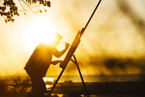 Silhouet van een jonge vrouw schilderij een foto op doek op een ezel, meisje profiel met verfborstel en palet bezig met kunst op de natuur bij zonsondergang — Stockfoto