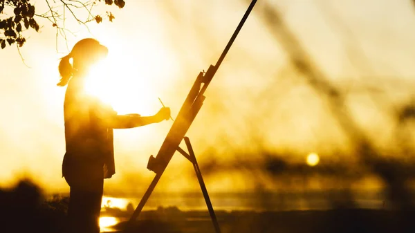 Silhouette di una donna che dipinge un quadro con vernici su tela su un cavalletto, ragazza con pennello e tavolozza impegnata in arte sulla natura in un campo al tramonto — Foto Stock