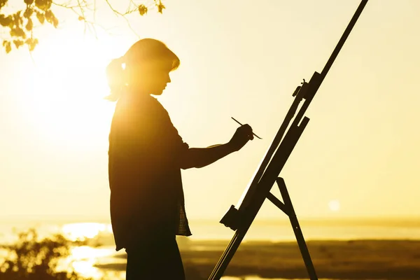 silhouette of a woman painting a picture with paints on canvas on an easel, girl with paint brush and palette engaged in art on the nature in a field at sunset