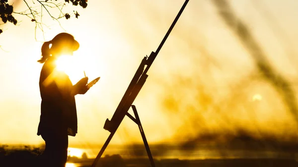 Silhouette di una donna che dipinge un quadro con vernici su tela su un cavalletto, ragazza con pennello e tavolozza impegnata in arte sulla natura in un campo al tramonto — Foto Stock