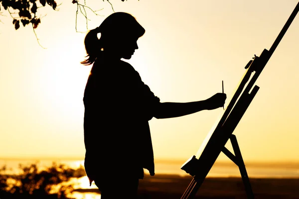 Silhueta de uma mulher pintando um quadro com pinturas em tela em um cavalete, menina com pincel de pintura e paleta envolvida em arte sobre a natureza em um campo ao pôr do sol — Fotografia de Stock