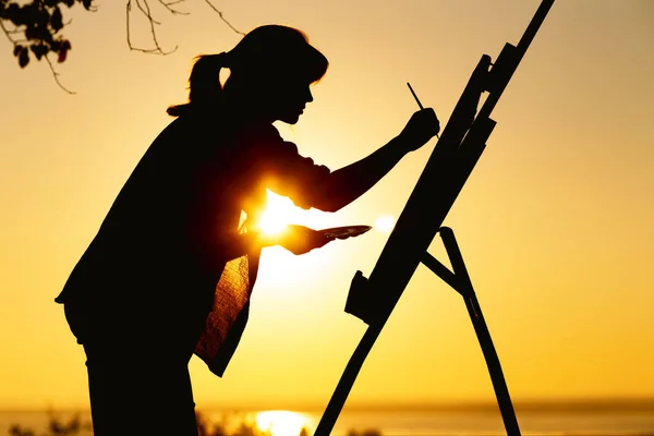 Silhouette einer Frau beim Bemalen eines Bildes mit Farben auf Leinwand auf einer Staffelei, Mädchen mit Pinsel und Palette in der Kunst auf einem Feld bei Sonnenuntergang — Stockfoto