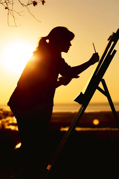 Silueta de una mujer pintando un cuadro con pinturas sobre lienzo sobre un caballete, niña con pincel y paleta dedicada al arte sobre la naturaleza en un campo al atardecer — Foto de Stock