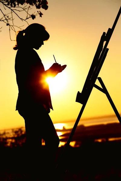 Silhouette einer jungen Frau, die ein Bild auf einer Staffelei in der Natur zeichnet, eine Frau, die mit Pinsel und Malerpalette unter dem Baum steht und sich bei Sonnenuntergang mit Kunst beschäftigt — Stockfoto