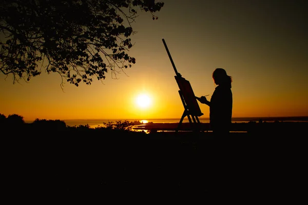 silhouette of a woman painting a picture with paints on canvas on an easel, girl with paint brush and palette engaged in art on the nature in a field at sunset