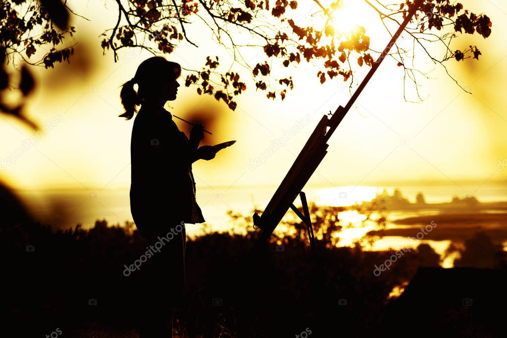 silhouette of a young woman drawing a picture on an easel on nature, female standing under tree with brush and artist's palette engaged in art at sunset