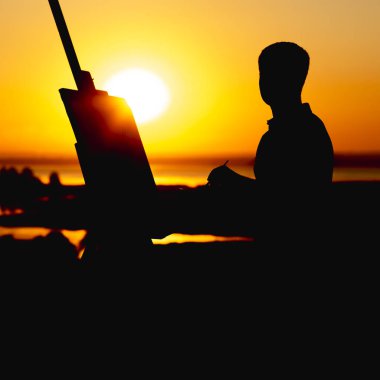 bir adamın silueti doğa üzerinde bir şelon üzerine tuval üzerine boyalar ile bir resim boyama, boya fırçası ve palet ile çocuk gün batımında sanat yapan