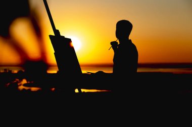 bir adamın silueti doğa üzerinde bir şelon üzerine tuval üzerine boyalar ile bir resim boyama, boya fırçası ve palet ile çocuk gün batımında sanat yapan