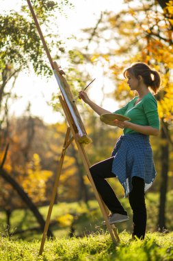 Tabloya resim çizen bir kadın doğada bir sehpaya, fırçalı ve paletli bir kız güzel manzarada bir resim, bir hobi konsepti