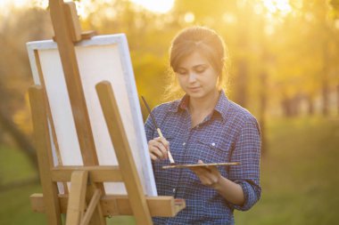 Kadın sanatçı doğadaki sehpaya resim çiziyor, fırçası ve paleti olan bir kız, yaratıcılık ve hobi kavramı