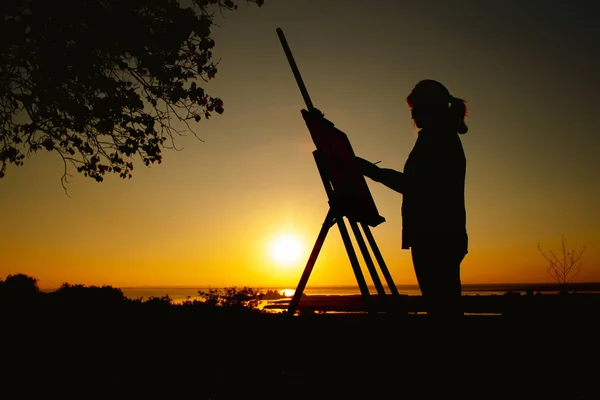 Silhueta de uma mulher pintando um quadro com pinturas em tela em um cavalete, menina com pincel de pintura e paleta envolvida em arte sobre a natureza em um campo ao pôr do sol — Fotografia de Stock