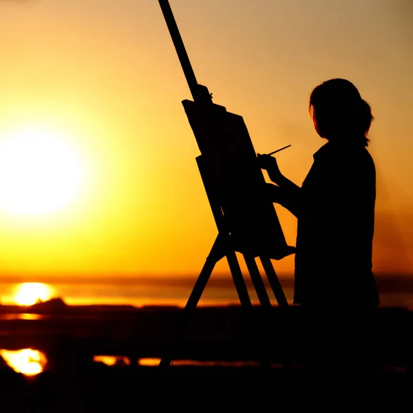 Силуэт молодой женщины, рисующей картину на мольберте на природе, девушка с кистью и палитрой художника, занимающейся искусством на закате солнца на горизонте — стоковое фото