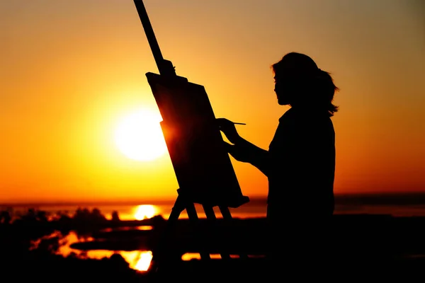 Силуэт молодой женщины, рисующей картину на мольберте на природе, девушка с кистью и палитрой художника, занимающейся искусством на закате солнца на горизонте — стоковое фото