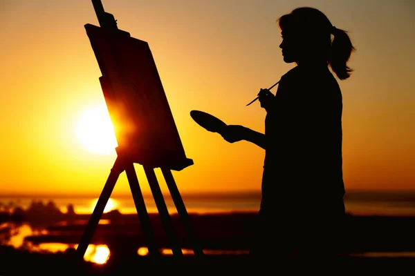 silhouette of a woman painting a picture with paints on canvas on an easel, girl with paint brush and palette engaged in art on the nature in a field at sunset