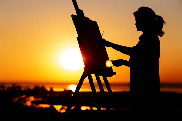Sziluettje egy nő festmény egy képet festékek vásznon egy festőállvány, lány ecset és a paletta foglalkozó művészeti jellegű területen a naplementekor — Stock Fotó
