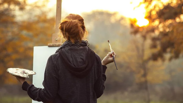 Rothaariges Mädchen mit einem Bild auf einer Staffelei in der Natur, eine junge Frau, die sich kreativ betätigt und die schöne Landschaft bei Sonnenuntergang genießt — Stockfoto
