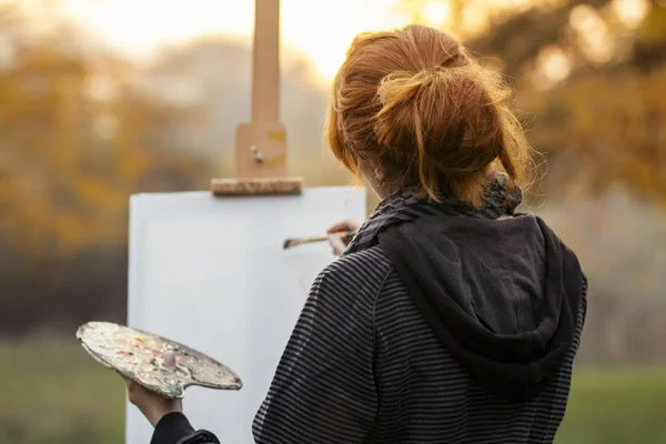 red-haired girl with painting a picture on an easel in nature, a young woman involved in creativity and enjoying beautiful landscape at sunset
