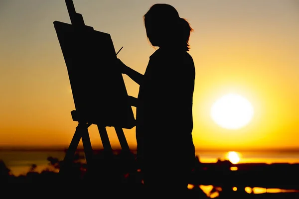 silhouette of a woman painting a picture with paints on canvas on an easel, girl with paint brush and palette engaged in art on the nature in a field at sunset