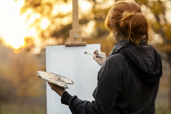 Doğada bir şelon üzerinde bir resim boyama ile kızıl saçlı kız, yaratıcılık katılan genç bir kadın ve gün batımında güzel manzara zevk — Stok fotoğraf