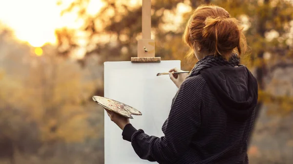Vörös hajú lány festmény egy képet egy festőállvány a természetben, egy fiatal nő részt vesz a kreativitás és élvezi gyönyörű táj naplementekor — Stock Fotó