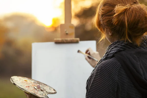 Chica pelirroja pintando un cuadro en un caballete en la naturaleza, una joven involucrada en la creatividad y disfrutando del paisaje — Foto de Stock