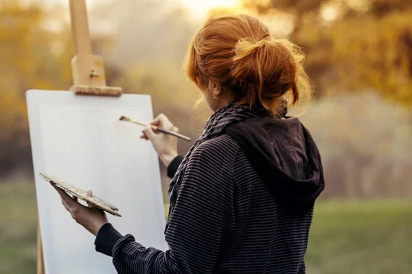 Fille aux cheveux roux avec la peinture d'un tableau sur un chevalet dans la nature, une jeune femme impliquée dans la créativité et profiter de beaux paysages au coucher du soleil — Photo