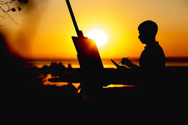 Silhueta de um jovem pintando um quadro em linho em um cavalete ao ar livre, perfil de cara de pintor envolvido na arte na natureza em um campo no pôr do sol — Fotografia de Stock