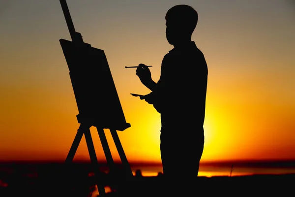 Силуэт мужчины, рисующего картину красками на холсте на мольберте на природе, мальчика с кистью и палитрой, занимающегося искусством на закате — стоковое фото
