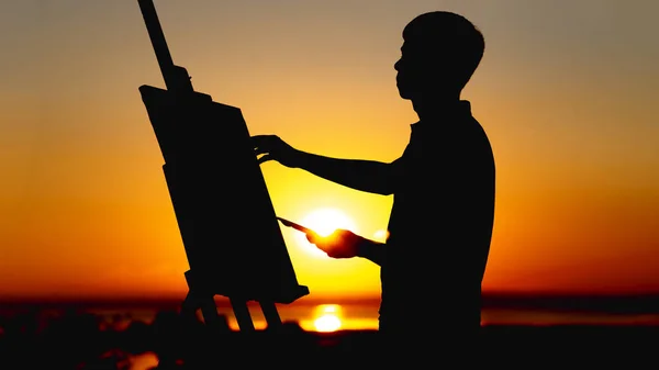 Силуэт человека, рисующего картину красками на холсте на мольберте на природе, парня с кистью и палитрой, занимающегося искусством на закате — стоковое фото