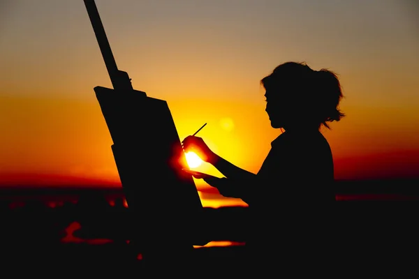 Силуэт женщины, рисующей картину на холсте на мольберте, девушки с кистью и палитрой, занимающейся искусством в поле на закате — стоковое фото